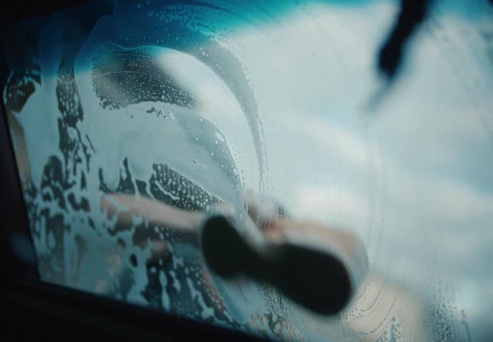 Slow motion interior view of a young man's face appearing through the soapy suds as he washes a car windshield with a sponge