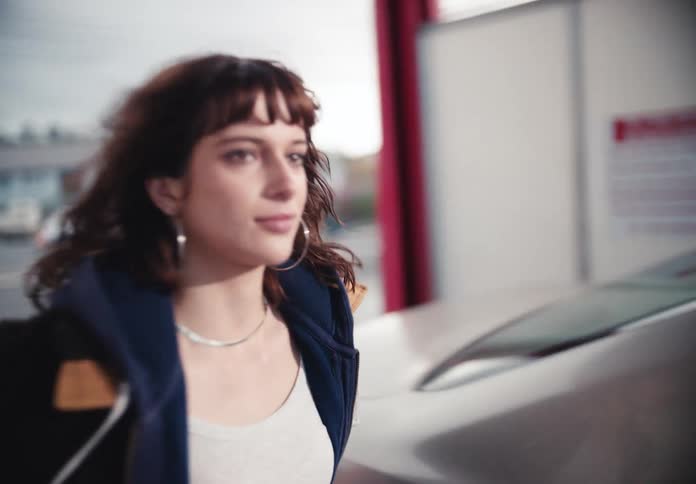 A cinematic close-up follows a young woman with dark hair and bangs walking in slow motion at an urban car wash