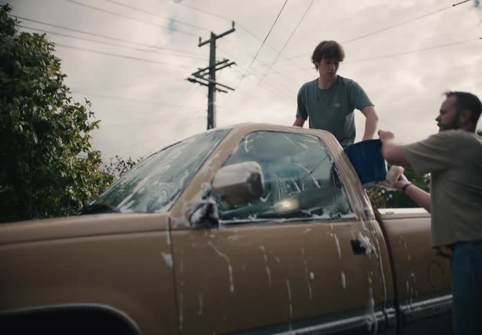 A father and his teenage son team up to wash their old pickup truck in the driveway on an overcast day
