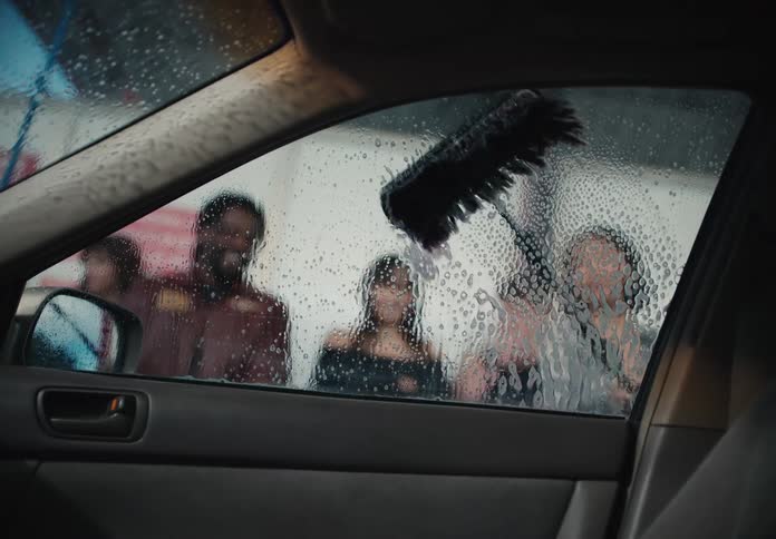 Workers wash a car window with a soapy brush, seen from the interior perspective in slow motion