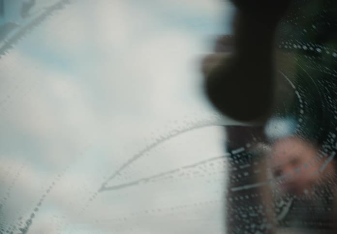An out-of-focus view from inside a car shows a hand wiping soapy suds off the windshield with a sponge