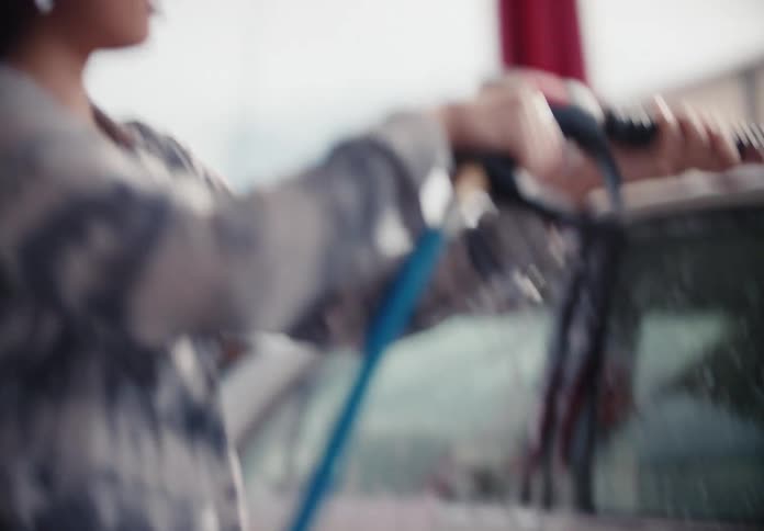A young woman in slow motion sprays her car clean with a pressure washer at a self-service car wash