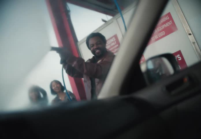 Smiling man playfully sprays a car windshield at a self-service car wash with his friends in slow motion, seen from inside the vehicle