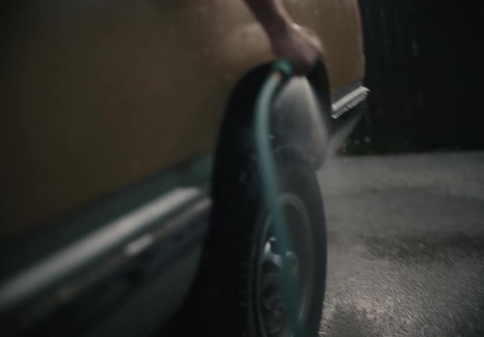 A young man in the back of a vintage pickup truck leans over to wash the tires with a hose on a cloudy day
