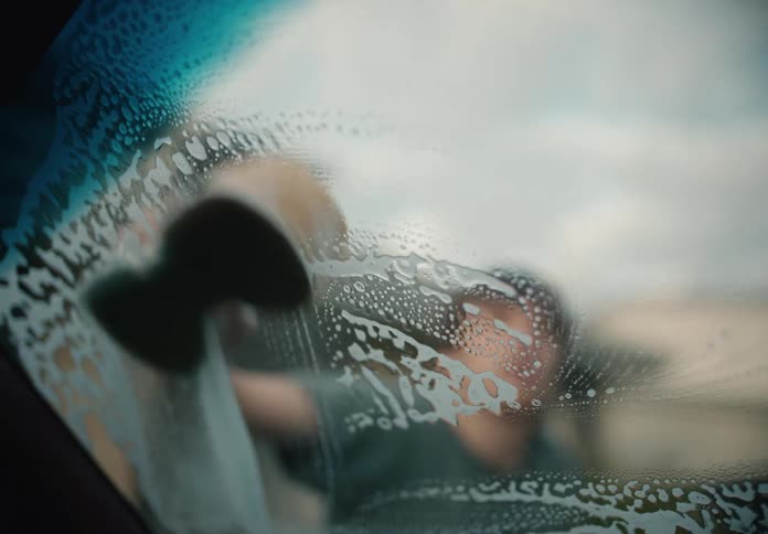 A young man washes a soapy car windshield with a sponge, filmed in slow motion from inside the vehicle