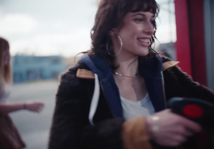 A young woman with dark hair laughs as she uses a pressure washer at a car wash with her friends in slow motion