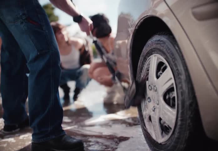 Low angle slow motion shot of a happy young man washing a car's tire with a soapy brush as his friends watch