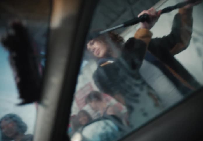 A gritty, cinematic interior shot of a young woman washing a car's windshield with a squeegee