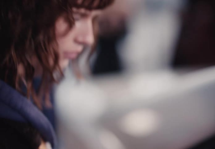 A contemplative young woman with brown wavy hair and bangs looks down in a thoughtful moment, captured in an intimate slow-motion close-up