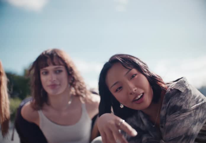 A trendy young Asian woman animatedly talks and gestures directly to the camera while hanging out with her friends on a sunny day