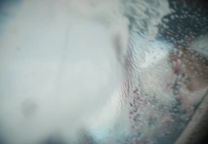 An abstract close-up from inside a car shows soapy water being sprayed and wiped off the windshield in slow motion