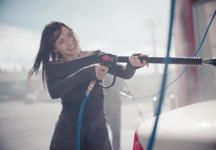 A stylish young woman playfully sprays down a white car with a high-pressure hose at a self-service car wash in slow motion