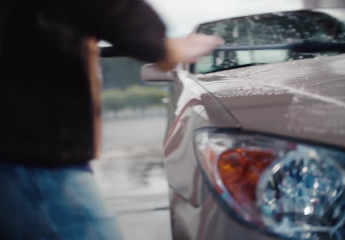 A person wearing a fur coat and hoodie scrubs the hood of a sedan with a brush at a car wash