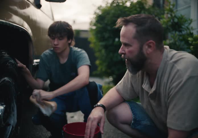 A father watches and instructs his teenage son on how to wash the car's tire in their driveway