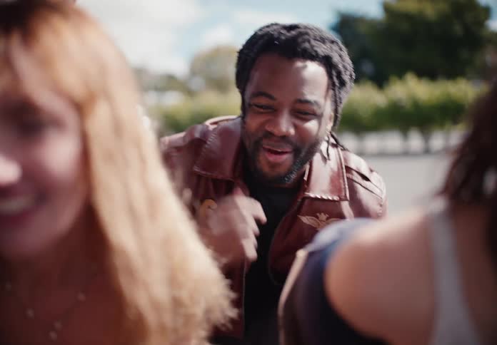 A charismatic young man in a brown leather jacket shares a joyful laugh with friends outdoors in slow motion