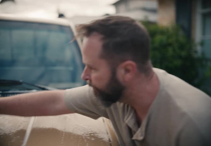 A middle-aged man with a beard washes his beige truck in the driveway, scrubbing the hood with a soapy sponge and then rinsing it with a hose