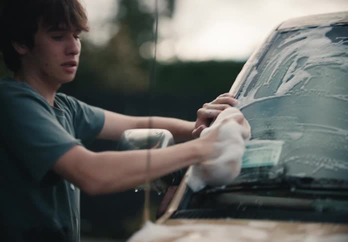 A young man carefully washes the windshield and hood of a car with a soapy sponge in slow motion