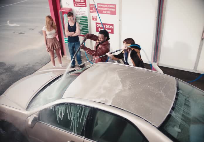 A high-angle slow-motion shot shows a diverse group of young friends playfully washing a car together at a self-service car wash