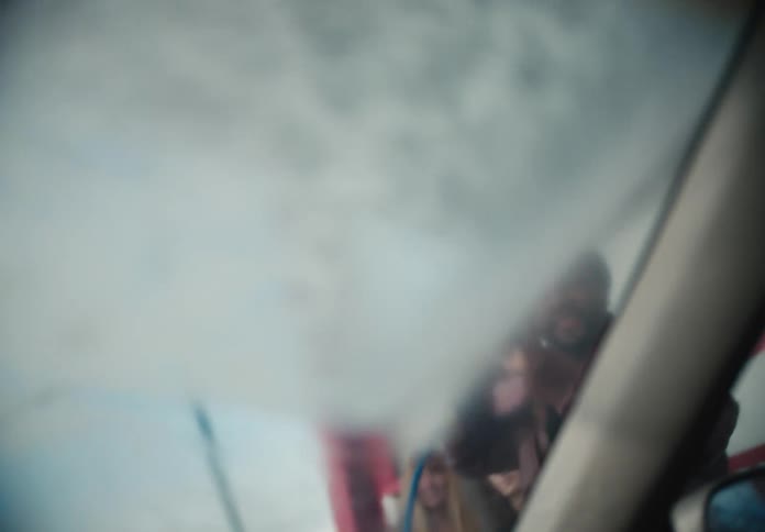 Playful man smiles while spraying a car windshield with a pressure washer, seen from inside the vehicle in slow motion
