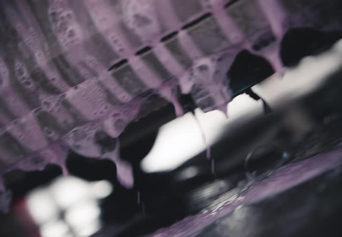 An abstract close-up shot captures purple soapy foam slowly dripping from a car's undercarriage in a car wash