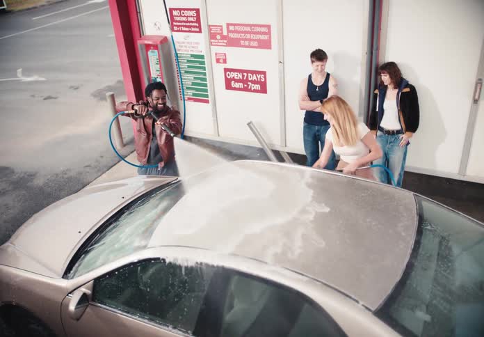 A diverse group of young friends have fun washing a car together at a self-service car wash in slow motion