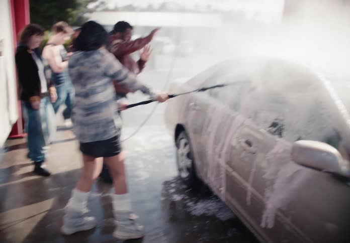 A group of diverse friends have fun at a self-service car wash, with one woman washing the car while another playfully sprays water and a man dances in slow motion