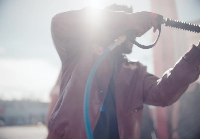 A smiling young man in a leather jacket playfully sprays a high-pressure water hose at a self-service car wash in slow motion