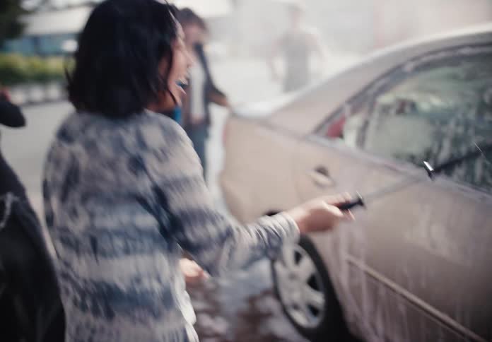 Happy Asian woman and her diverse friends dance and laugh while washing a car at a self-service bay in slow motion