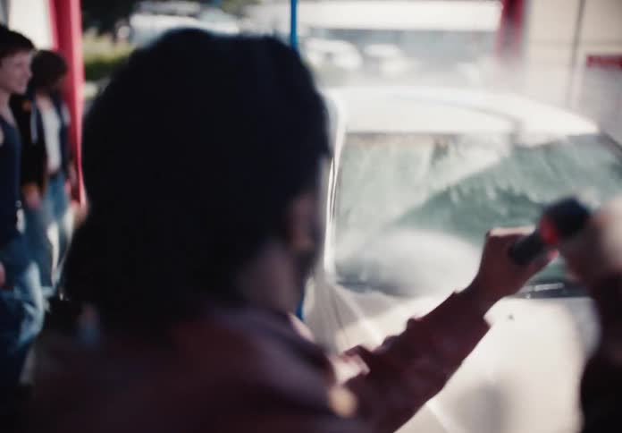 Young man takes over washing a car from his friend with a high-pressure water hose at a car wash in slow motion
