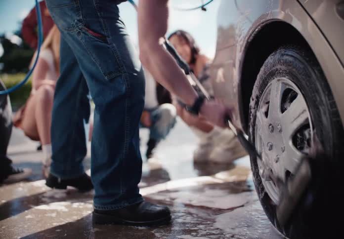 A young man vigorously scrubs his car's tire at a self-service car wash as his friends watch in the background