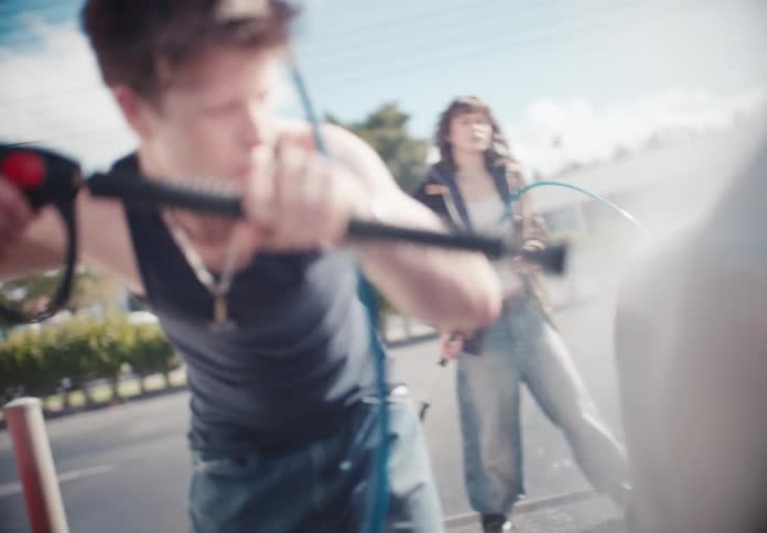 A smiling young man in a tank top playfully sprays a car with a high-pressure hose at an outdoor car wash