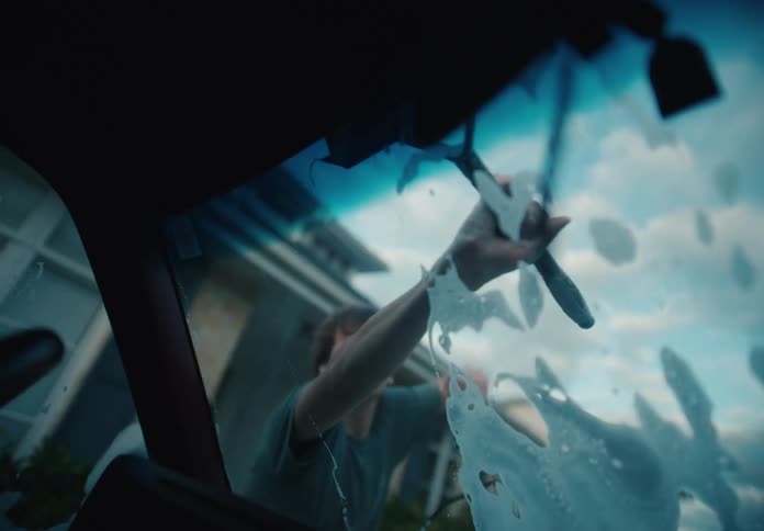 Slow motion footage from inside a car shows a young man washing the soapy windshield with a squeegee as his father helps