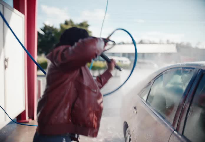 A smiling man joyfully dances while washing his car with a pressure washer at a self-service bay in slow motion