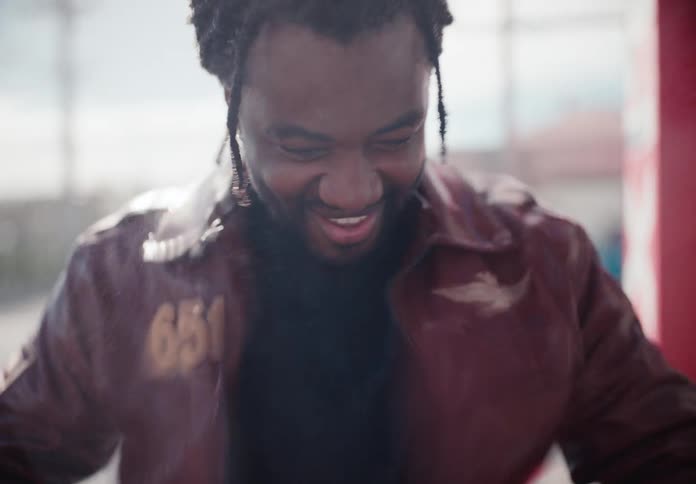 A cheerful man in a leather jacket laughs playfully as he's sprayed with water in slow motion on a sunny day