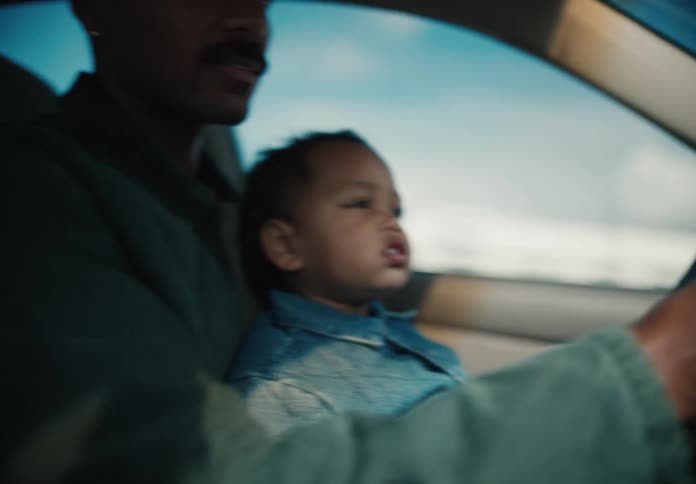 A toddler sits on his father's lap and holds the steering wheel as they drive together on a sunny day
