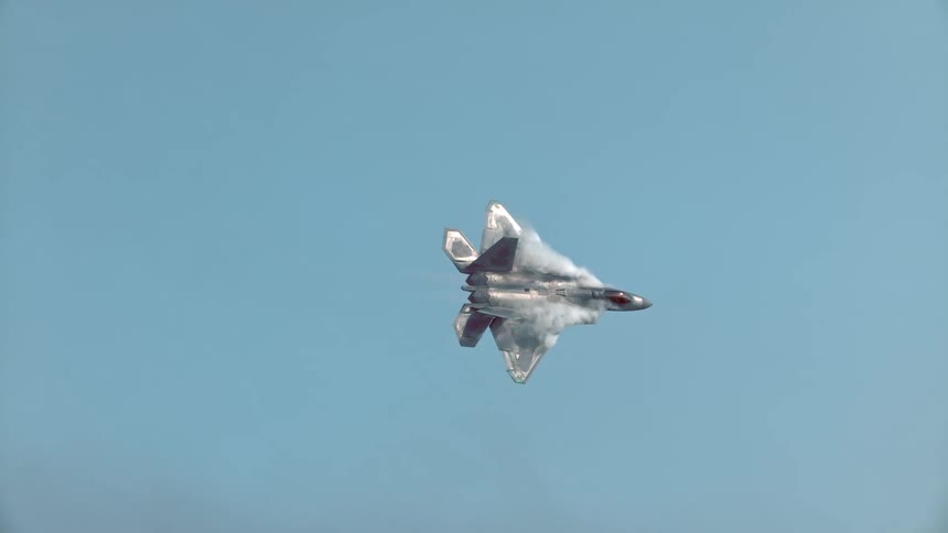 An F-22 Raptor fighter jet soars through the blue sky in slow motion, generating dramatic vapor trails before vanishing into the clouds