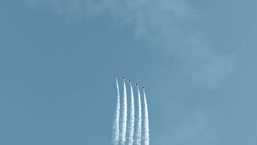 Five aerobatic jets perform a synchronized maneuver, soaring upwards and leaving long white smoke trails against the clear blue sky