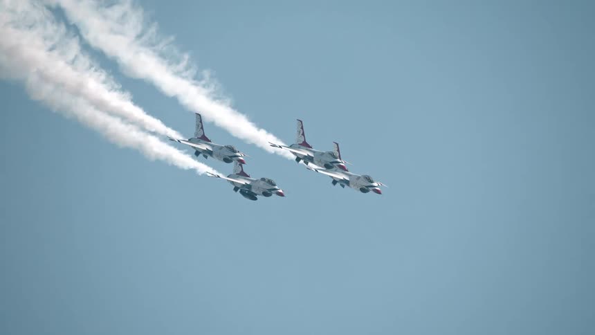 A squadron of four fighter jets performs a synchronized flight in slow motion, leaving long white smoke trails against the vast blue sky