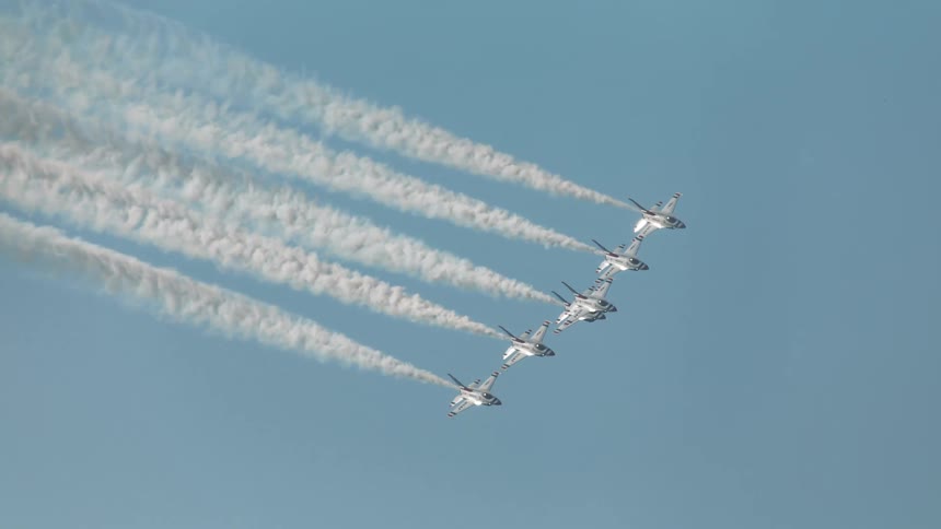 A squadron of five fighter jets performs an aerobatic maneuver in slow motion, leaving smoke trails across the clear blue sky