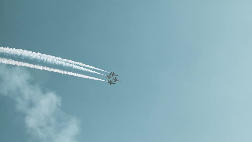 Four fighter jets fly in a precise formation, leaving long white smoke trails across the sky in slow motion