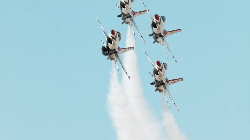 Four USAF Thunderbirds fighter jets fly in a tight diamond formation, releasing white smoke in a slow-motion climb against a clear blue sky