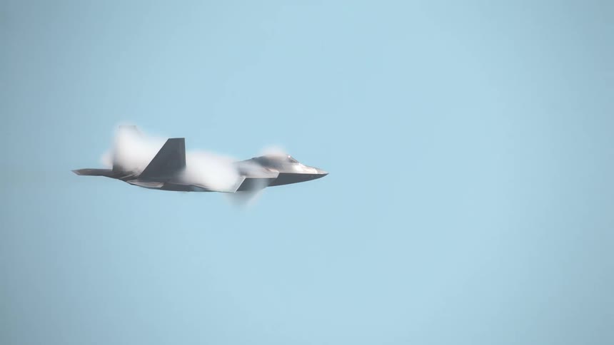 An F-22 Raptor fighter jet flies in slow motion, creating a stunning vapor cone as it approaches transonic speed against a clear blue sky