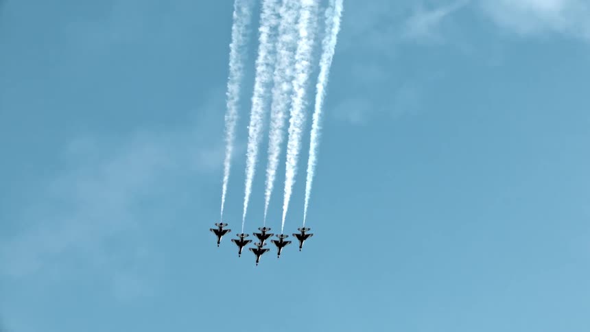 A squadron of six aerobatic jets ascends in tight formation, leaving long white smoke trails against a pale blue sky