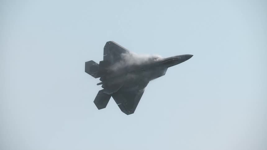 An F-22 Raptor fighter jet performs a high-alpha maneuver, creating vapor over its wings as it flies in slow motion through a hazy sky