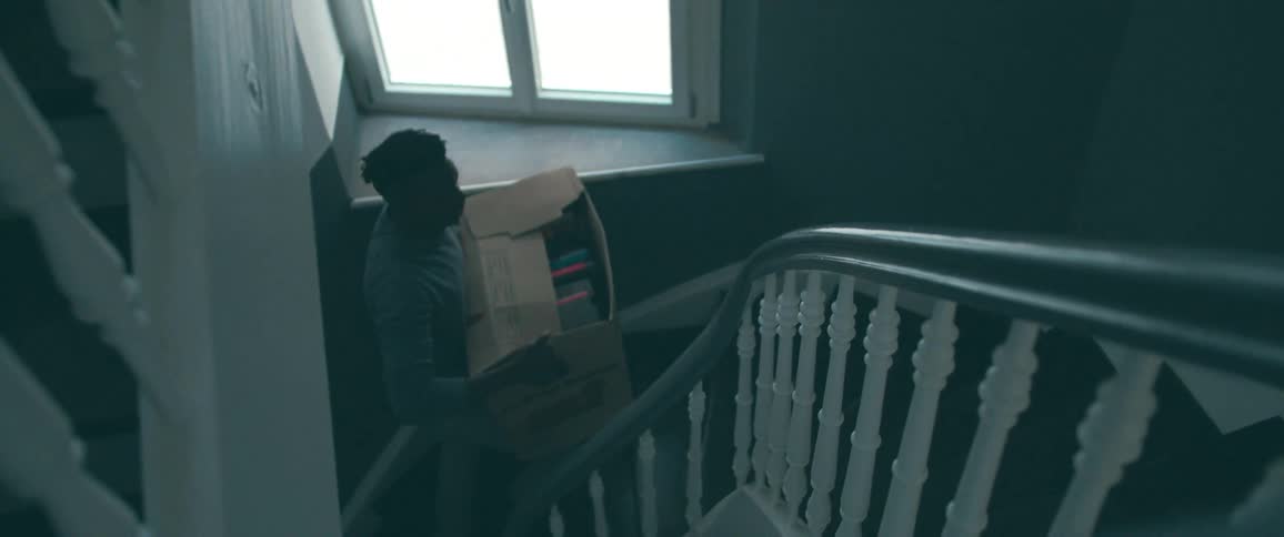 High-angle slow-motion shot of a man carefully carrying a heavy box of belongings down a winding staircase as he moves house