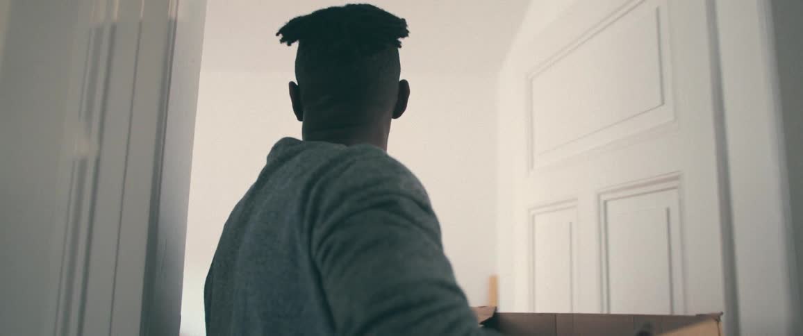 A man seen from behind walks in slow motion through a hallway carrying a moving box into a bright, empty room