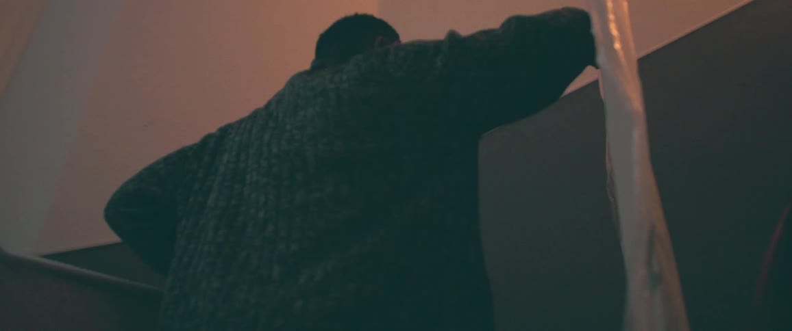 A low-angle, slow-motion shot shows a man in a dark sweater leaning against the wall of a dimly lit stairwell
