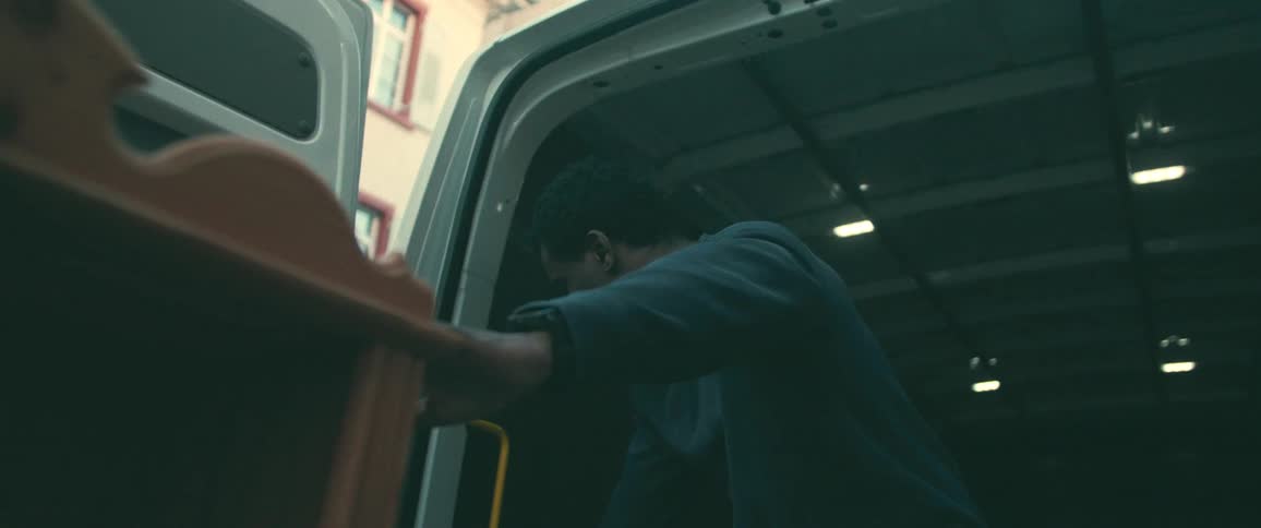 A young man in slow motion carefully unloads wooden furniture pieces from the back of a white delivery van