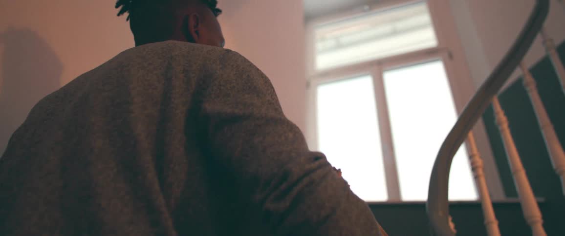 A cinematic low angle shot from behind captures a man in a sweater walking up an indoor staircase towards a bright window