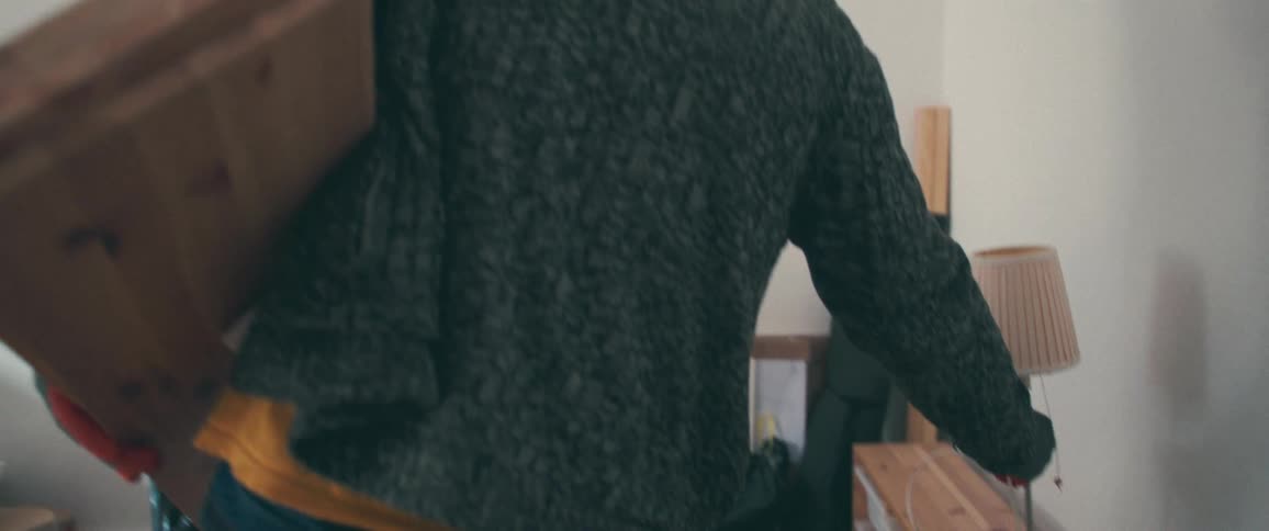 Slow motion shot of a man's back as he carries moving boxes and a lamp to place them in a cluttered room